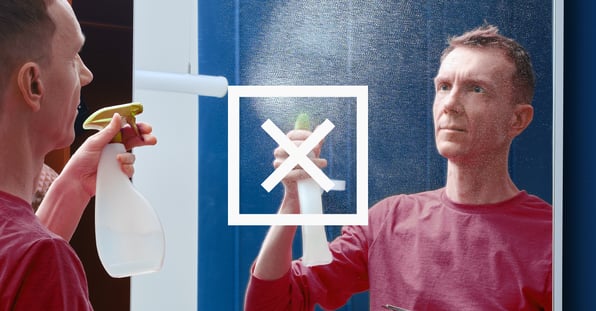 A man cleaning a mirror incorrectly, by spraying cleaner directly on the surface instead of on a soft cloth.